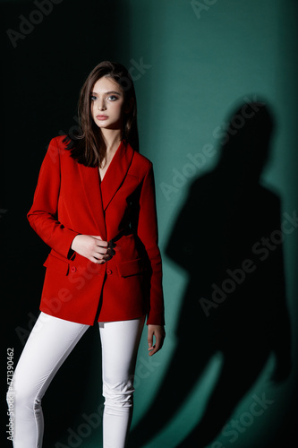 Fashion model in red jacket and white trousers, pants posing in deep shadow on emerald, spruce green color background in a studio.