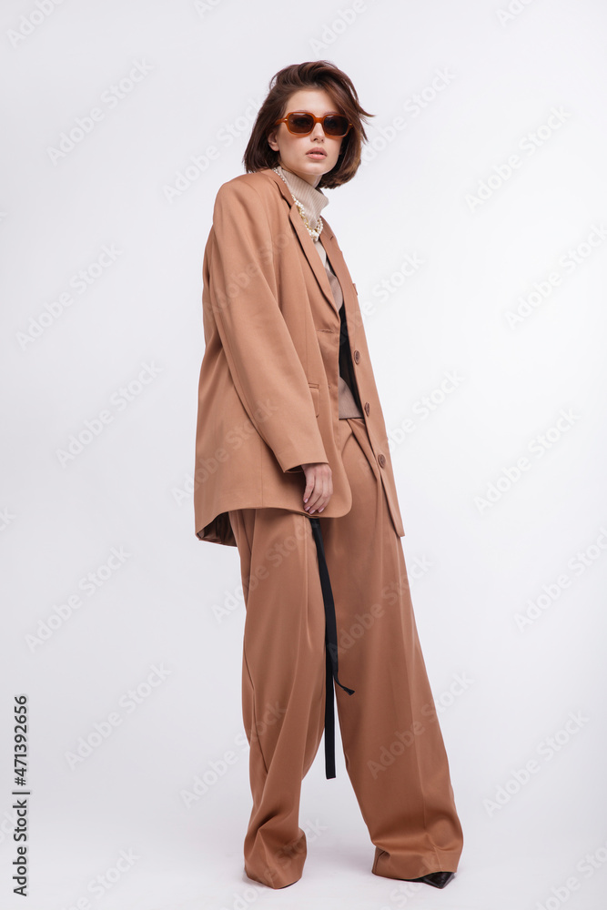 Fashion photo of a beautiful elegant young woman in a pretty oversize brown and beige suit, corset, stylish sunglasses, jacket, pants posing over white background. Bob haircut. Studio Shot. Portrait Fashion photo of a beautiful elegant young woman in a pretty oversize brown and beige suit, corset, stylish sunglasses, jacket, pants posing over white background. Bob haircut. Studio Shot. Portrait