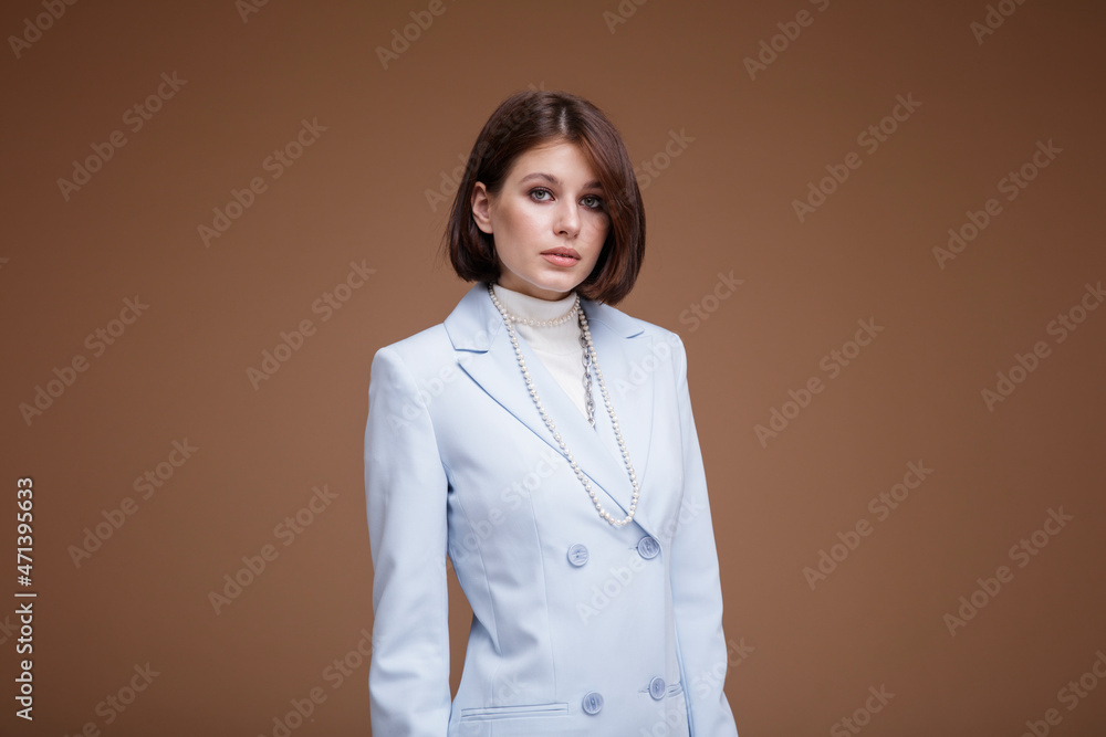 Fashion photo of a beautiful elegant young woman in a pretty blue suit in the style of a flight attendant stewardess, pearl necklace posing over brown background. Bob haircut. Studio Shot. Fashion photo of a beautiful elegant young woman in a pretty blue suit in the style of a flight attendant stewardess, pearl necklace posing over brown background. Bob haircut. Studio Shot.