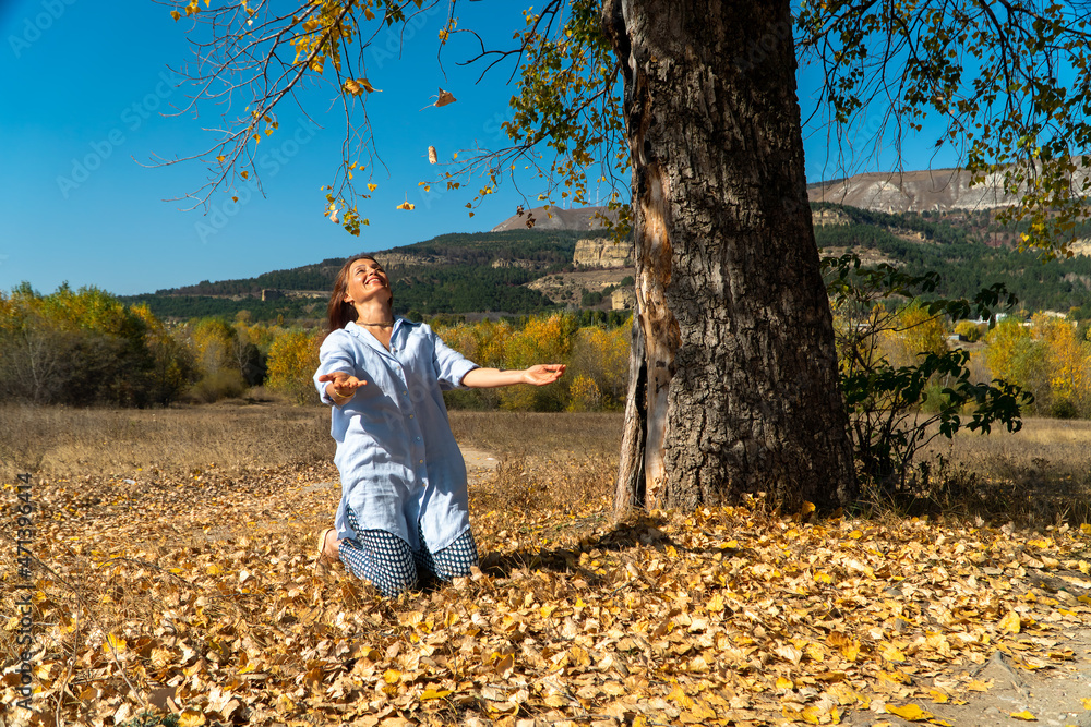 Smiling woman in blue long shirt sitting under a tree and tossing up autumn leaves