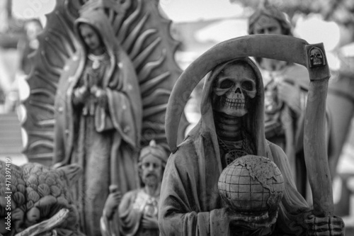 Black and White Close Up View of Santa Muerte Grim Reaper Statue and Virgin Mary Figures for Sale at Street Market Outdoor Public Square, Mexican Folk Religious Tradition Art Culture Background
