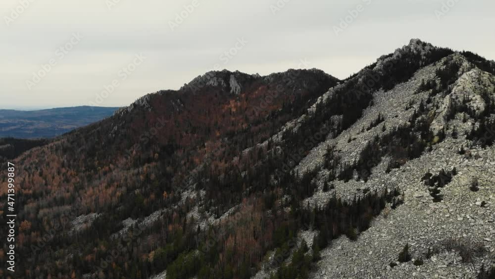 Aerial view of the top of the mountain from pile of stones in the autumn forest. Flying over green and gray trees. Other mountain peaks can be seen in the distance. Late autumn. Drone shooting Aerial view of the top of the mountain from pile of stones in the autumn forest. Flying over green and gray trees. Other mountain peaks can be seen in the distance. Late autumn. Drone shooting