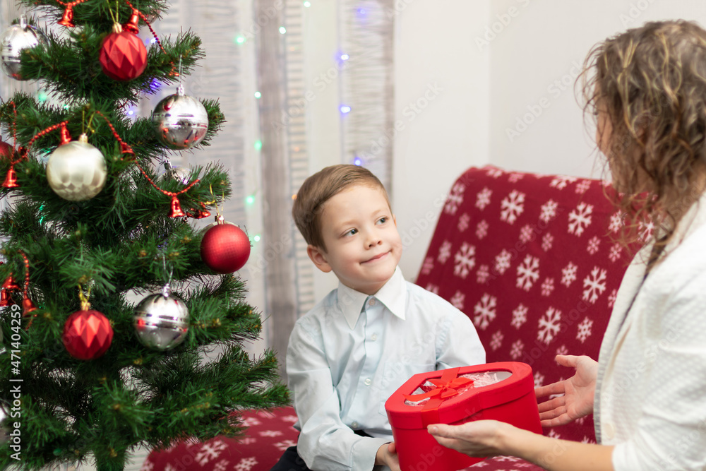A seven-year-old boy in a smart shirt gives his mother a gift in a box for the New Year against the backdrop of a Christmas tree at home. Selective focus. Portrait A seven-year-old boy in a smart shirt gives his mother a gift in a box for the New Year against the backdrop of a Christmas tree at home. Selective focus. Portrait