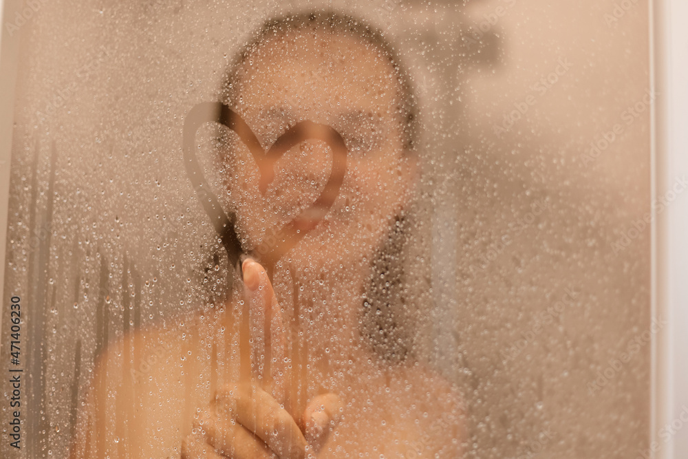Photo of smiling attractive young woman draws heart on weeping glass shower door, enjoys rest in douche, washing her body, standing behind steam blurred glass with water drops. - obrazy, fototapety, plakaty 