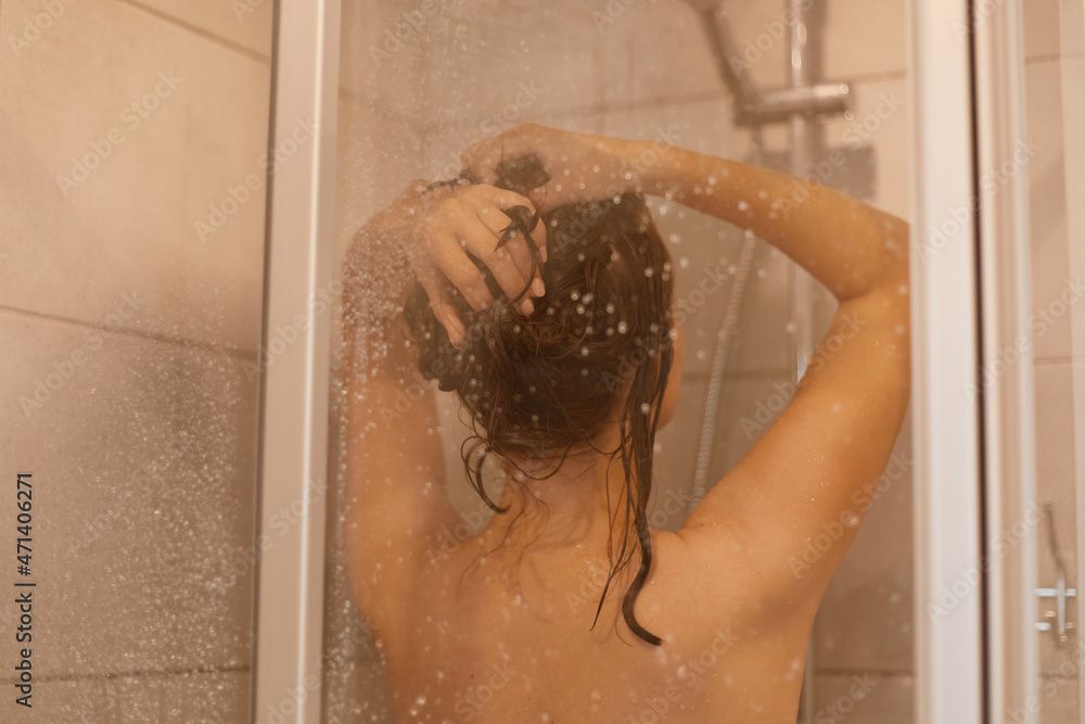 Side view shot of young woman with perfect naked body standing with raised arms under water, enjoying taking shower, washing her hair, doing hygienic procedures.