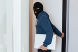 Portrait of man robber wearing blue hoodie and black balaclava standing with stolen laptop, looking at camera, opens door to leave the house he robbed.
