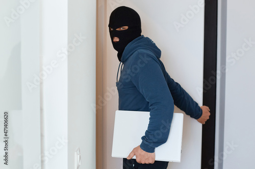 Portrait of man robber wearing blue hoodie and black balaclava standing with stolen laptop, looking at camera, opens door to leave the house he robbed.