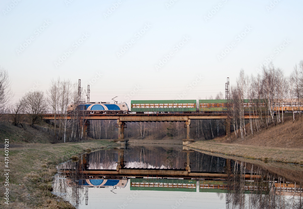 View of a small river in the spring against the background of a passing train with freight cars on a railway bridge. Channel of the Vileyka-Minsk water system for the transfer of water flow