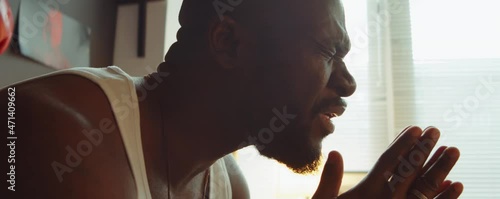 Close up tilt down shot of African American man sitting with eyes closed, holding hands clasped and saying prayer at home
