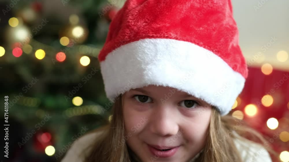 Close up Portrait of happy surprised little girl wearing Santa hat on background of decorated Christmas tree and New Year lights. Child Looks at Camera with delight and surprise at face. Wow Emotion Close up Portrait of happy surprised little girl wearing Santa hat on background of decorated Christmas tree and New Year lights. Child Looks at Camera with delight and surprise at face. Wow Emotion