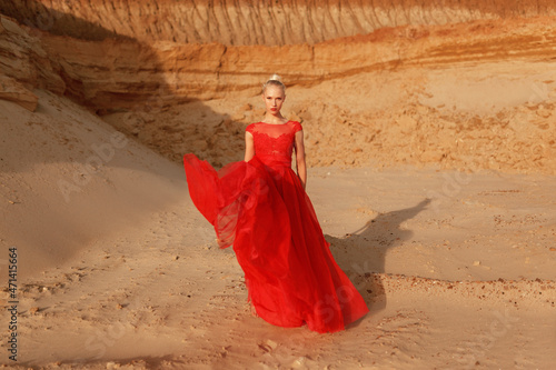 Blonde woman in red waving dress with flying fabric on the background of golden sands.