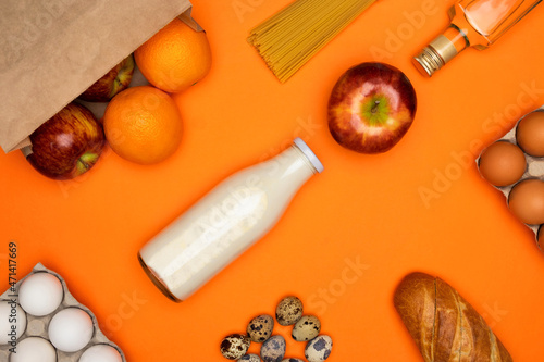 Red apples and oranges in paper eco bag. Spaghetti, bottle of milk, eggs, bread, olive oil on orange background. Healthy food, delivery, donation concept. Food stock for quarantine. Top view, flatly.