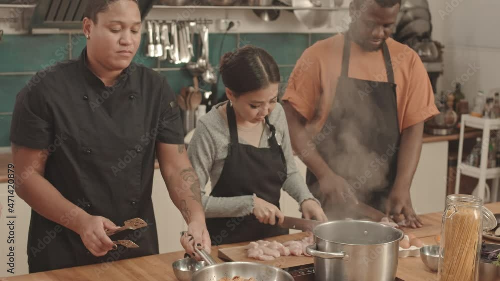 Medium of young female chef roasting chicken in pan on stove, Asian woman and African American man standing by table in large kitchen, cutting fillets, Caucasian sous-chef watching Medium of young female chef roasting chicken in pan on stove, Asian woman and African American man standing by table in large kitchen, cutting fillets, Caucasian sous-chef watching
