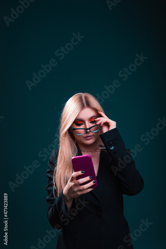 Young caucasian business woman using a mobile phone wearing a black blazer and glasses in a photography studio with a blue dark background.