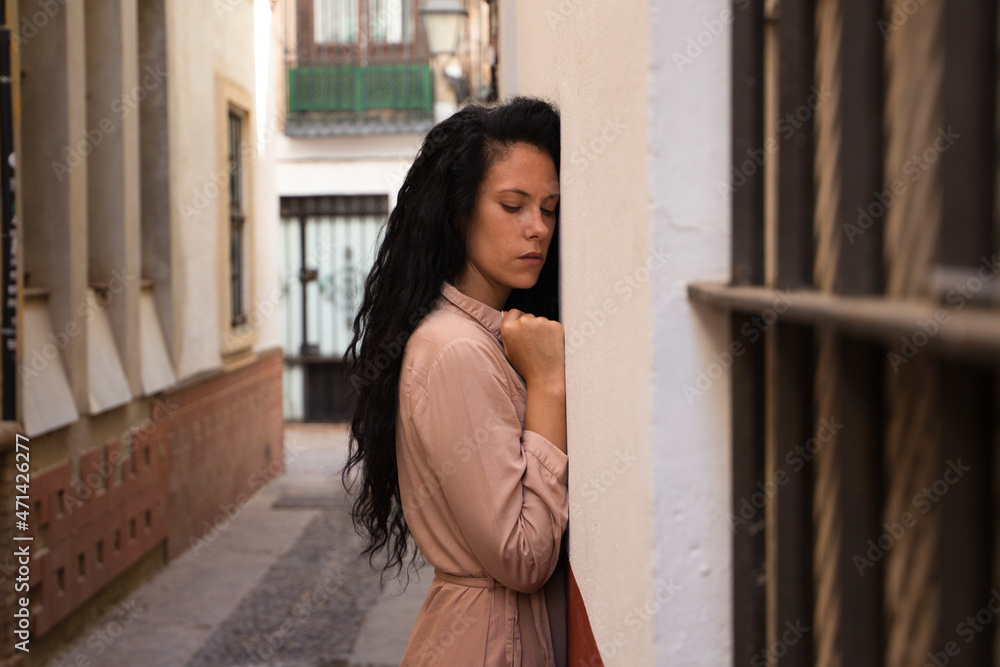 beautiful woman with dark curly hair leaning against the wall of a typical european house in a mediterranean style city. The woman is serious and sad. Concept expressions.