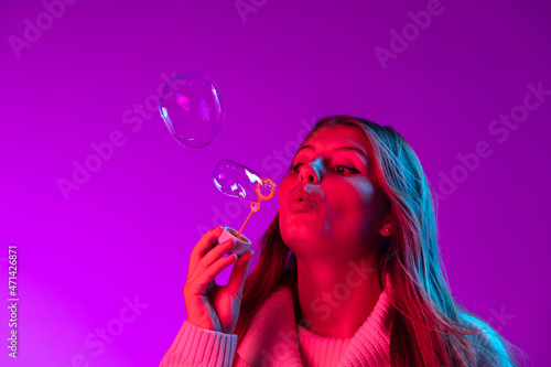 Young beautiful girl with cute freckles in white sweater blowing soap bubbles isolated on purple color studio background in neon light.