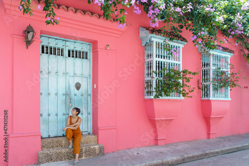 Side view of woman sit on a pink wall in Cartagena de Indias. Horizontal view of latin woman sightseeing in spanish historic ancient city. Travel to Colombia concept.