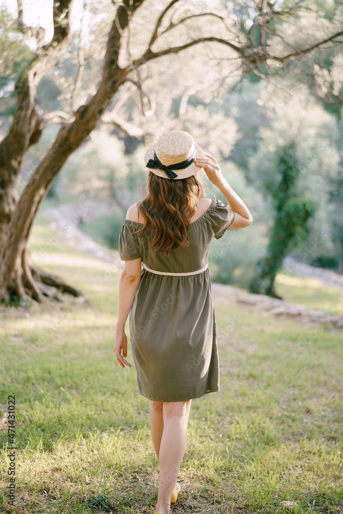 Girl in a green dress and hat walks along a green lawn under a tree. Back view