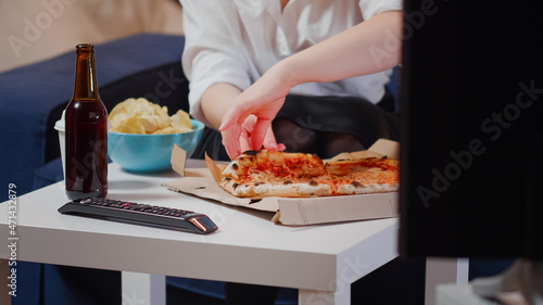 Close up of white table with unhealthy food and beer in living room. Young woman eating slice of pizza from delivery box sitting on couch at television. Person with takeaway meal at home