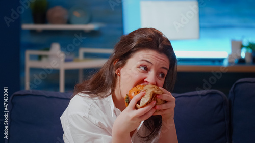 Caucasian woman taking bite from hamburger in living room and looking at television. Young adult eating fast food takeout with beer for dinner while sitting on couch with movie on TV