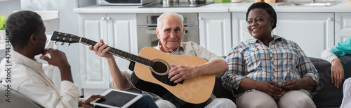 Senior man playing acoustic guitar near multiethnic friends with digital tablet in nursing home, banner