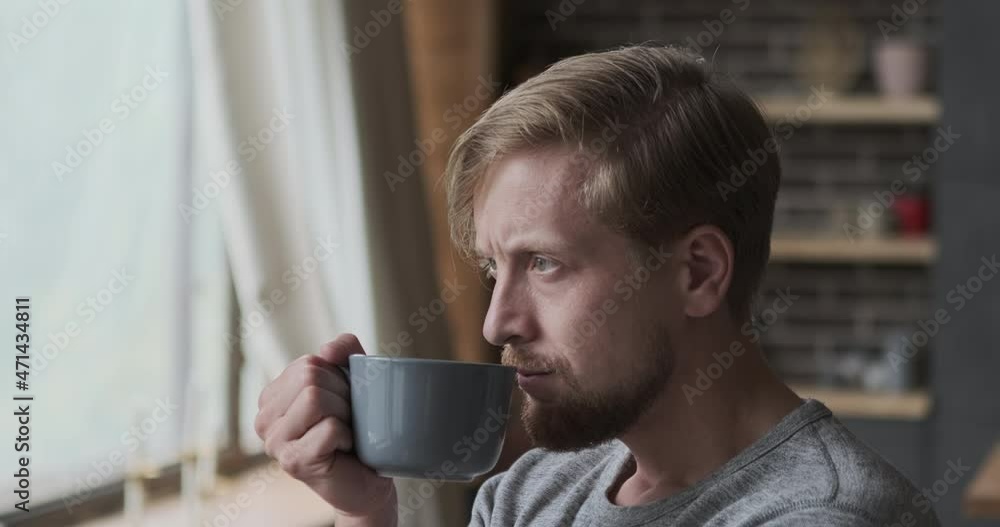 Pensive stylish man dreaming thinking about future drinking hot beverage from mug close up. Wistful blonde male contemplating street through window enjoying morning tea coffee or having break at home Pensive stylish man dreaming thinking about future drinking hot beverage from mug close up. Wistful blonde male contemplating street through window enjoying morning tea coffee or having break at home