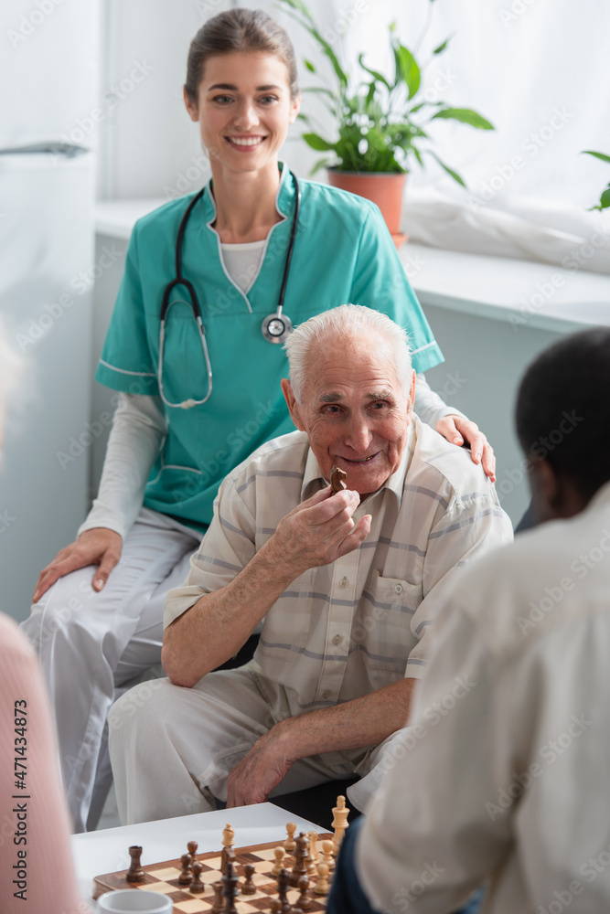 Obraz premium Senior man holding chess figure near smiling nurse and african american friend in nursing home
