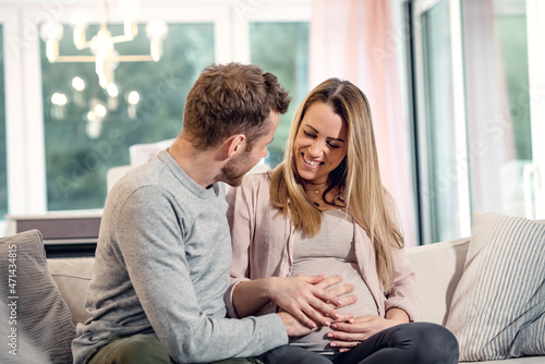 Young pregnant happy couple sitting on couch husband stroking baby belly of blond wife