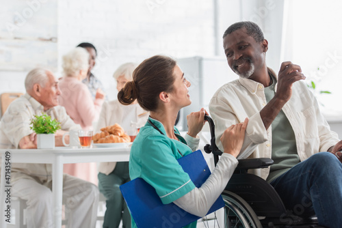Cheerful nurse with clipboard talking with african american man in wheelchair in nursing home photo
