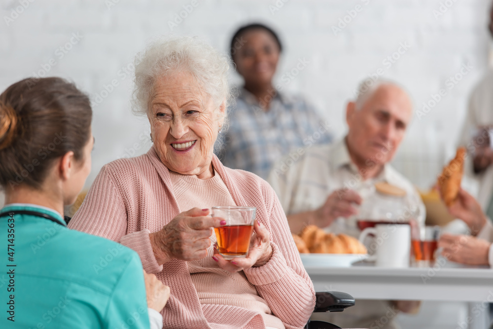 Positive senior woman holding tea while talking to blurred nurse in nursing home