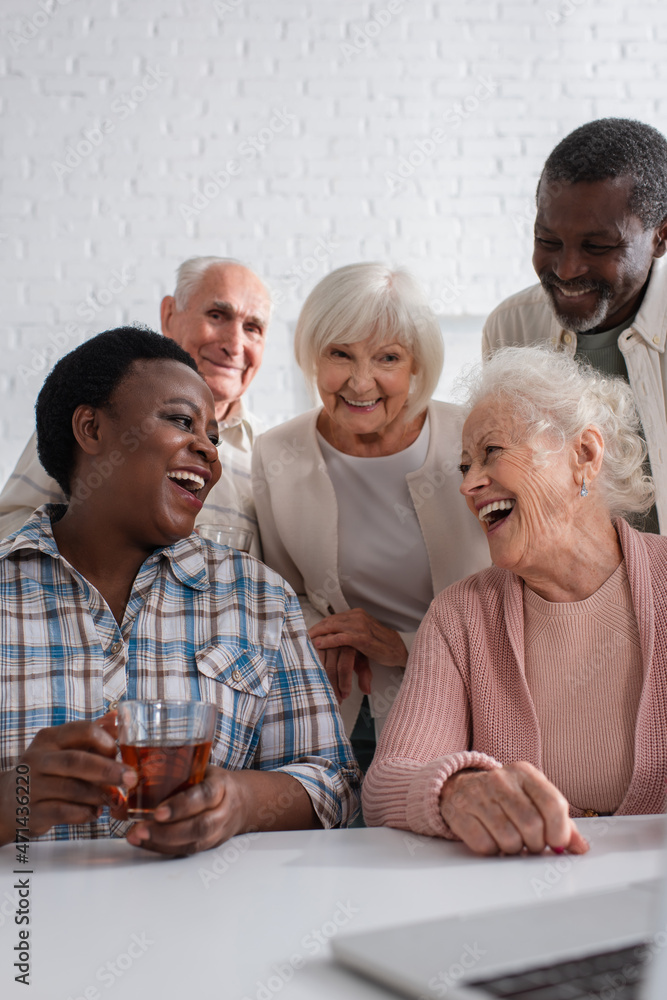 African american woman holding tea near smiling friends and laptop in nursing home African american woman holding tea near smiling friends and laptop in nursing home