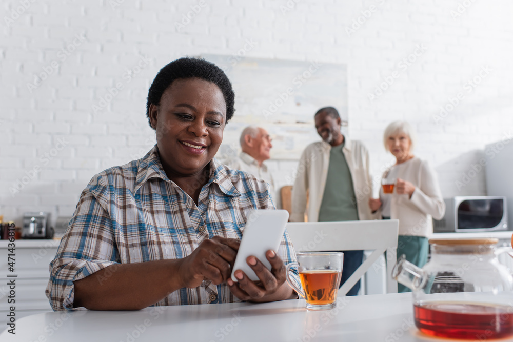 Smiling african american woman using smartphone near tea and blurred people in nursing home Smiling african american woman using smartphone near tea and blurred people in nursing home
