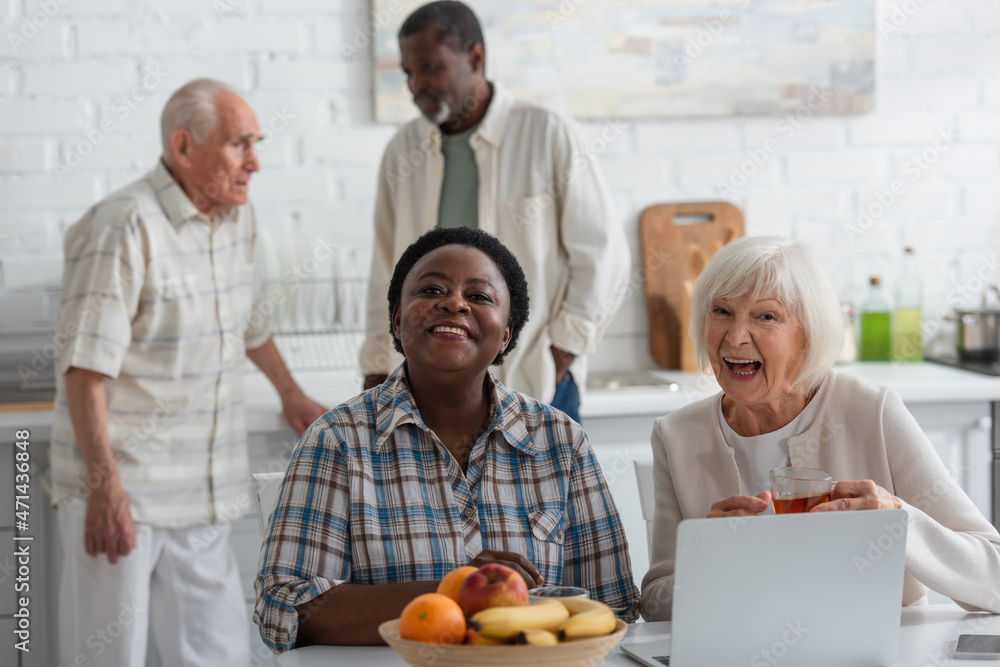 Cheerful multiethnic women with tea looking at camera near devices and fruits in nursing home Cheerful multiethnic women with tea looking at camera near devices and fruits in nursing home