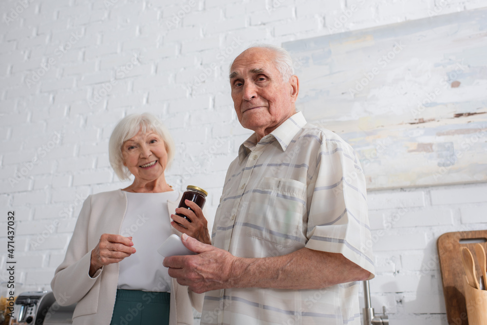 Low angle view of senior man holding smartphone near friend with jar of jam in nursing home Low angle view of senior man holding smartphone near friend with jar of jam in nursing home