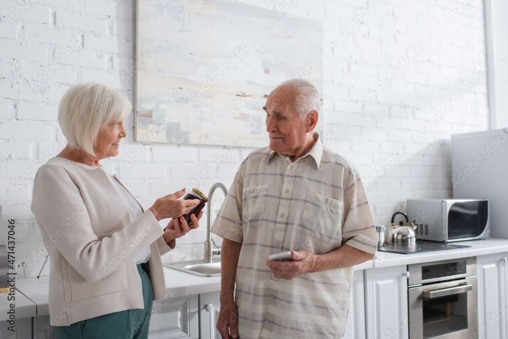 Obraz premium Smiling senior woman pointing at jar with jam near friend with smartphone in nursing home
