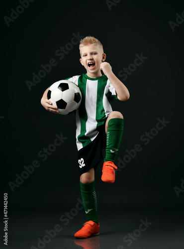 Young soccer player  teenager boy in red white striped uniform celebrates win  goal  stands holding ball and gesturing over black background