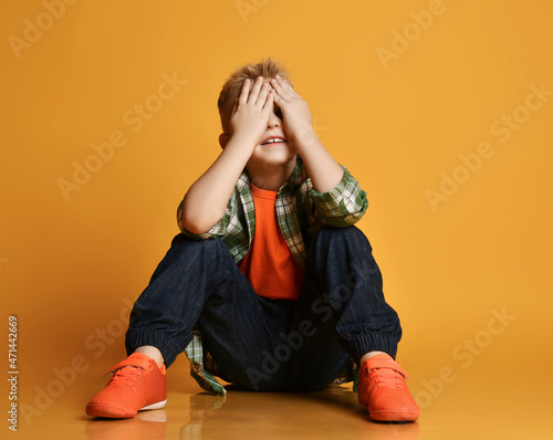 Blond smiling boy child in checkered shirt and sneakers sitting and covering eyes with hands over yellow background. Trendy casual children fashion, fantasy concept photo