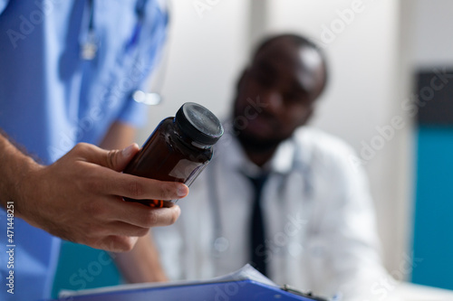 Closeup of caucasian man nurse holding pills bottle in hand analyzing medicine prescription working in hospital office. African american doctor explaining medical treatment. Healthcare service
