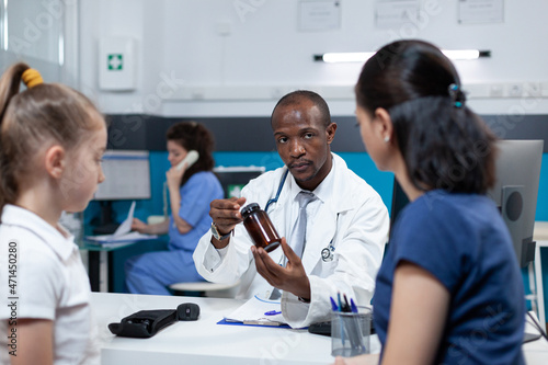 African american pediatrician doctor discussing disease symptoms explaining antibiotics treatment holding vitamin bottle during clinical inspection in hospital office. Health care service