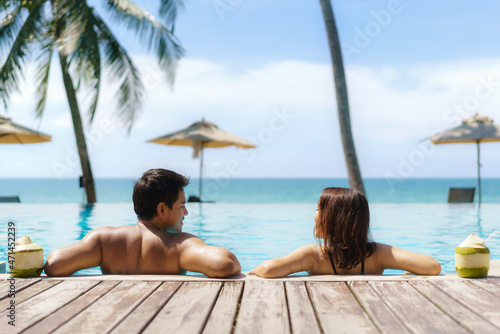 Summer travel vacation concept, traveler asian couple with coconut relax in luxury infinity pool hotel resort with sea beach and palm tree background