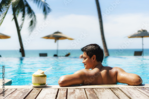 Summer travel vacation concept, Traveler asian man with coconut relax in luxury infinity pool hotel resort with sea beach and palm tree background