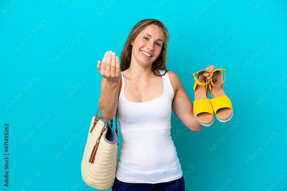 Young caucasian woman holding summer sandals isolated on blue background inviting to come with hand. Happy that you came Young caucasian woman holding summer sandals isolated on blue background inviting to come with hand. Happy that you came