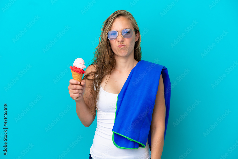 Young caucasian woman holding ice cream and wearing a beach towel isolated on blue background with sad expression Young caucasian woman holding ice cream and wearing a beach towel isolated on blue background with sad expression