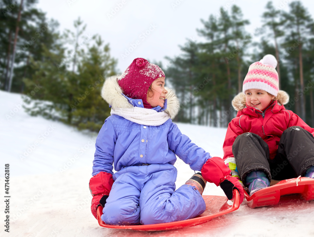 childhood, leisure and season concept - happy little girls on sleds outdoors in winter over snowy forest or park background childhood, leisure and season concept - happy little girls on sleds outdoors in winter over snowy forest or park background