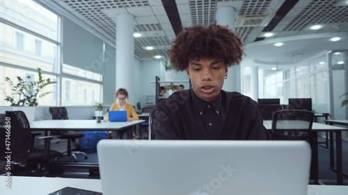 African American employee using laptop in office and mumbling something, diverse coworkers on background. Arc shot man working online, browsing internet. Concept of business