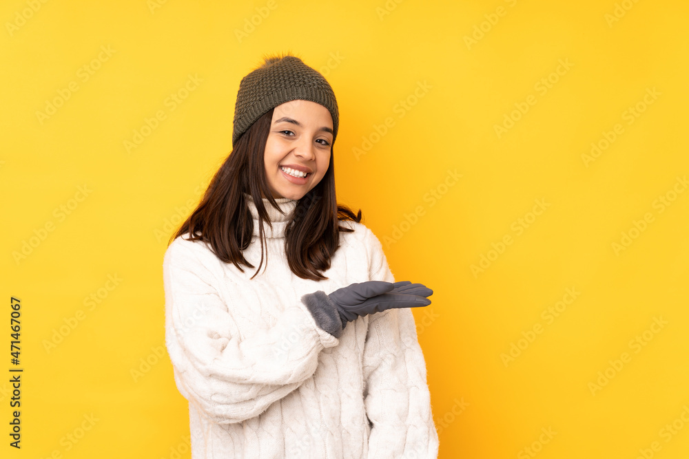 Young woman with winter hat over isolated yellow background presenting an idea while looking smiling towards Young woman with winter hat over isolated yellow background presenting an idea while looking smiling towards