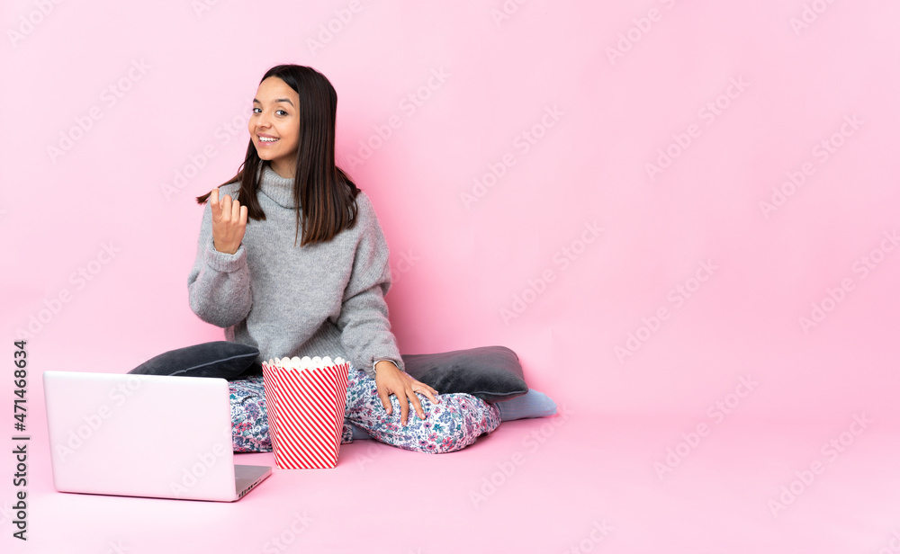 Young mixed race woman eating popcorn while watching a movie on the laptop doing coming gesture Young mixed race woman eating popcorn while watching a movie on the laptop doing coming gesture