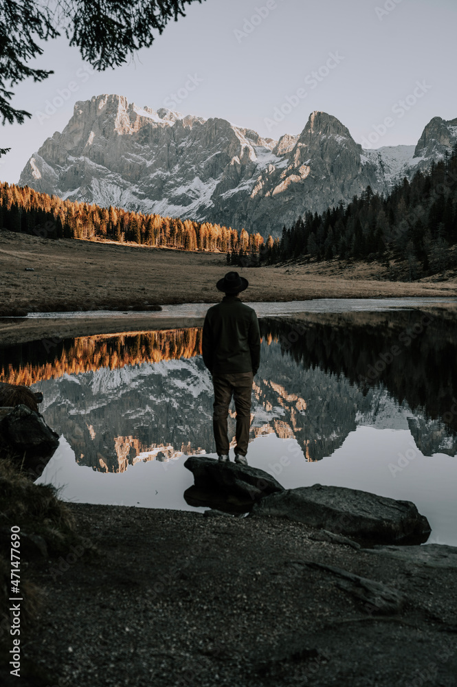 A man with a hat is standing by a mountain lake in the Dolomites. The mountains and the rest of the landscape are reflected in the lake. It is autumn. The Calaita lake