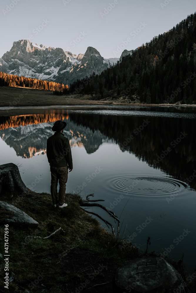 A man with a hat is standing by a mountain lake in the Dolomites. The mountains and the rest of the landscape are reflected in the lake. It is autumn. The Calaita lake another one