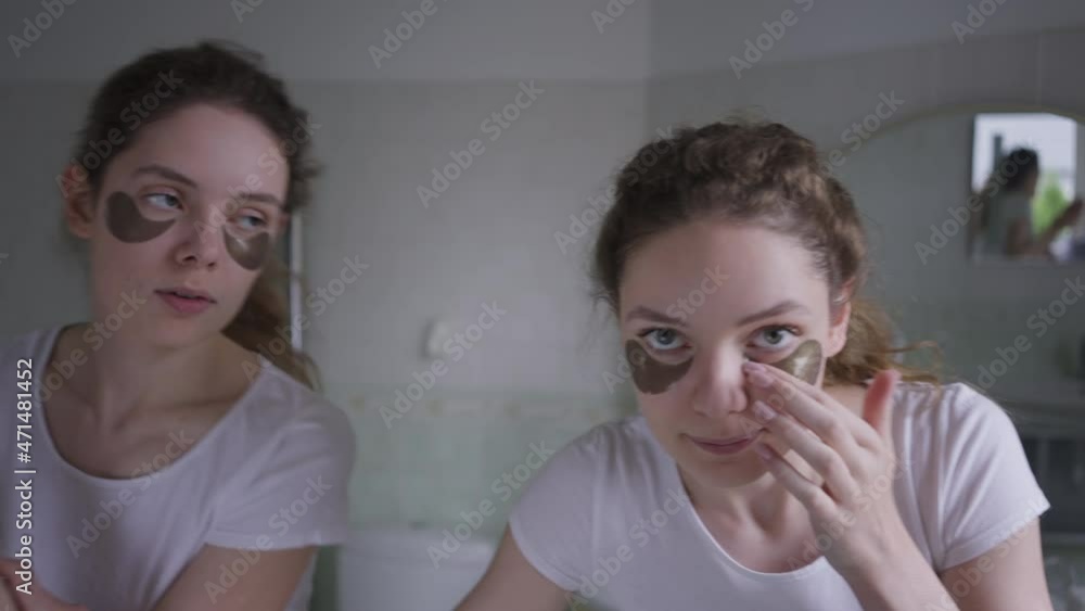 Front view portrait twin sisters fighting for mirror applying moisturizing eye mask in bathroom at home. Slim beautiful Caucasian young women taking care of facial skin in the morning. Beauty routine Front view portrait twin sisters fighting for mirror applying moisturizing eye mask in bathroom at home. Slim beautiful Caucasian young women taking care of facial skin in the morning. Beauty routine